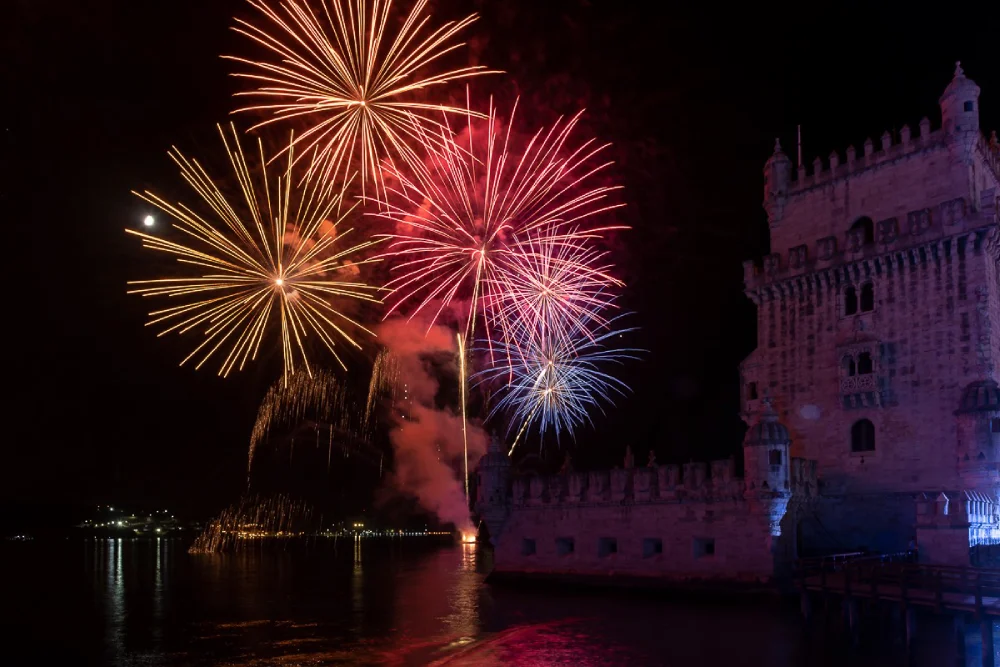 Con un meteo favorevole, nel cielo i fuochi d'artificio colorati scoppiano sulla torre di Belem vicino al fiume Tejo nella notte di Capodanno a Lisbona in Portogallo. Il cielo è illuminato da rossi, arancioni e blu brillanti, che si riflettono nell'acqua calma sottostante, creando una scena abbagliante.