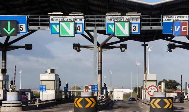 Viene mostrato un casello autostradale con diverse corsie, con cartelli chiari per i metodi di pagamento e le tariffe autostradali alla periferia di Lisbona. Le frecce verdi guidano i veicoli nelle corsie contrassegnate, comprese quelle riservate ai pendolari. Una struttura sul tetto copre i caselli sotto un cielo limpido.