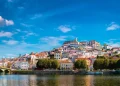 Una vista pittoresca di Coimbra, città da visitare, rivela una città storica in collina adornata da edifici colorati e un grande fiume che scorre in primo piano. Il cielo azzurro brillante è punteggiato di nuvole e un ponte abbellisce il lato sinistro mentre rigogliosi alberi verdi costeggiano la riva del fiume.