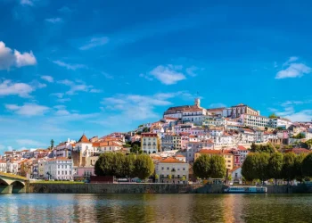 Una vista pittoresca di Coimbra, città da visitare, rivela una città storica in collina adornata da edifici colorati e un grande fiume che scorre in primo piano. Il cielo azzurro brillante è punteggiato di nuvole e un ponte abbellisce il lato sinistro mentre rigogliosi alberi verdi costeggiano la riva del fiume.
