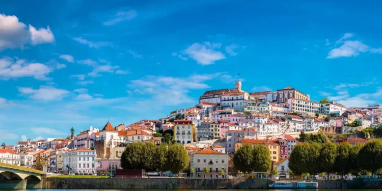 Una vista pittoresca di Coimbra, città da visitare, rivela una città storica in collina adornata da edifici colorati e un grande fiume che scorre in primo piano. Il cielo azzurro brillante è punteggiato di nuvole e un ponte abbellisce il lato sinistro mentre rigogliosi alberi verdi costeggiano la riva del fiume.