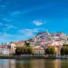 Una vista pittoresca di Coimbra, città da visitare, rivela una città storica in collina adornata da edifici colorati e un grande fiume che scorre in primo piano. Il cielo azzurro brillante è punteggiato di nuvole e un ponte abbellisce il lato sinistro mentre rigogliosi alberi verdi costeggiano la riva del fiume.