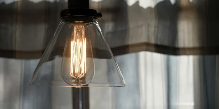 Una lampadina accesa all'interno di una casa EDP Comercial, design a sospensione in vetro trasparente è appesa davanti a tende trasparenti, proiettando una luce calda. Lo sfondo è caratterizzato da una luce naturale dolcemente diffusa attraverso le tende.