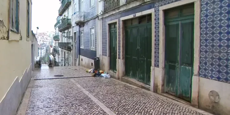 Una stretta strada acciottolata, dove è avvenuta a Lisbona la violenza di un italiana, fiancheggiata da vecchi edifici con piastrelle decorative e balconi in ferro battuto. Le porte verdi si affacciano sulla strada, con un paio di sacchi della spazzatura sul marciapiede. In discesa, in lontananza, che fondono magnificamente le culture.