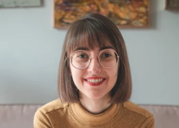 Marta Lamalfa, con i capelli castani lunghi fino alle spalle e gli occhiali, sorride calorosamente mentre è seduta all'interno di una libreria portoghese. Indossa elegantemente un maglione color senape. Dietro di lei, un dipinto delicatamente messo a fuoco adorna la parete di colore chiaro, aggiungendo all'atmosfera serena.