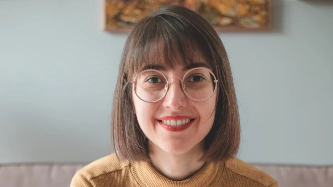 Marta Lamalfa, con i capelli castani lunghi fino alle spalle e gli occhiali, sorride calorosamente mentre è seduta all'interno di una libreria portoghese. Indossa elegantemente un maglione color senape. Dietro di lei, un dipinto delicatamente messo a fuoco adorna la parete di colore chiaro, aggiungendo all'atmosfera serena.