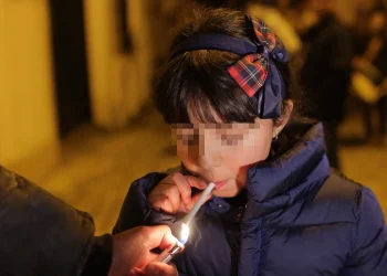 Durante il tradizionale rito in cui i bambini fumano in Portogallo, una ragazzina con un cappotto blu è in piedi con gli occhi chiusi mentre qualcuno le accende una sigaretta. Indossa una fascia per capelli con un fiocco scozzese. Lo sfondo è leggermente sfocato, suggerendo un'ambientazione notturna.