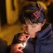 Durante il tradizionale rito in cui i bambini fumano in Portogallo, una ragazzina con un cappotto blu è in piedi con gli occhi chiusi mentre qualcuno le accende una sigaretta. Indossa una fascia per capelli con un fiocco scozzese. Lo sfondo è leggermente sfocato, suggerendo un'ambientazione notturna.