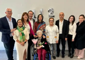 Un gruppo di persone è in piedi e sorride attorno a alla donna più vecchia del Portogallo. Palloncini argentati mostrano il numero "112" sul muro dietro di loro.
