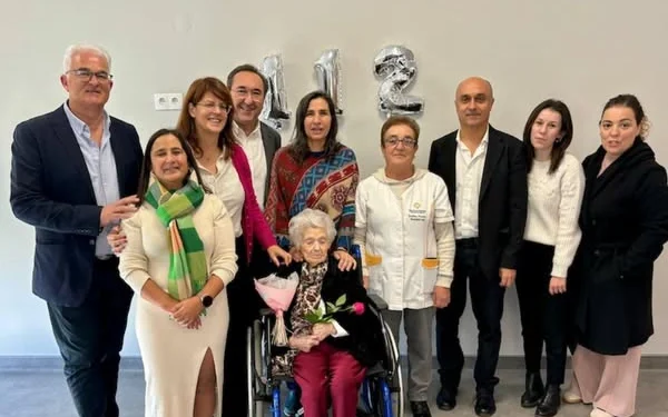 Un gruppo di persone è in piedi e sorride attorno a alla donna più vecchia del Portogallo. Palloncini argentati mostrano il numero "112" sul muro dietro di loro.