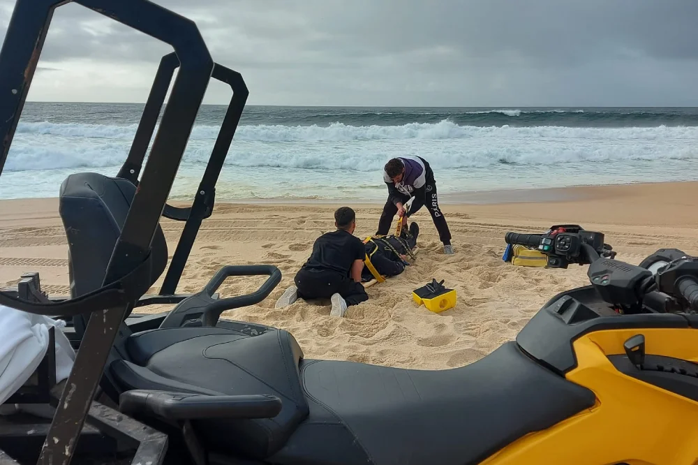 Su una spiaggia sabbiosa, due italiani salvano una donna dalle onde del mare. Nelle vicinanze ci sono attrezzature di salvataggio, una borsa gialla e un fuoristrada. Le onde dell'oceano si infrangono sullo sfondo sotto un cielo nuvoloso.