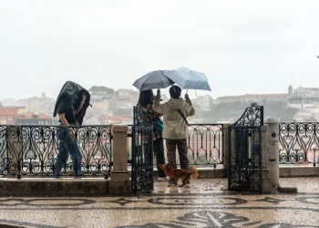 Tre persone e un cane sono in piedi su una terrazza di Lisbona che domina un paesaggio urbano, in una giornata con meteo piovoso. Due tengono l'ombrello mentre uno usa la giacca come riparo. La terrazza presenta ringhiere decorate e un pavimento piastrellato, con la città velata di nebbia sullo sfondo.