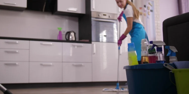 In costa una colf con un grembiule blu e guanti rosa lava il pavimento in una cucina moderna in Portogallo, quanto muovendosi in modo efficiente. Prodotti per la pulizia, tra cui un secchio con varie bottiglie, sono in primo piano. La cucina è caratterizzata da mobili bianchi e un tappeto sul pavimento.
