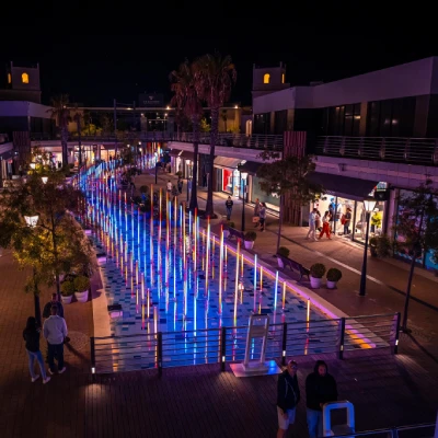Una vivace passerella per lo shopping all'aperto di notte, fiancheggiata da negozi, abbraccia l'eccitazione dei saldi in Portogallo. Barre luminose verticali illuminate mostrano colori blu e viola, creando un percorso colorato. Alberi e persone in primo piano contribuiscono all'atmosfera vivace.