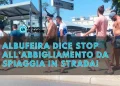 Un gruppo di persone in bikini cammina lungo una strada vicino a un cartello con la scritta Albufeira centro. Il testo in italiano sopra l'immagine recita: Albufeira dice stop all’abbigliamento da spiaggia in strada!.