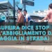 Un gruppo di persone in bikini cammina lungo una strada vicino a un cartello con la scritta Albufeira centro. Il testo in italiano sopra l'immagine recita: Albufeira dice stop all’abbigliamento da spiaggia in strada!.