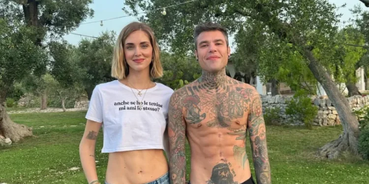 Chiara Ferragni con un crop top bianco e jeans è in piedi accanto a Fedez senza maglietta con tatuaggi in un'area erbosa con alberi, non in Portogallo, bensì sono in Puglia. Sorridono sotto le luci scintillanti, catturando l'essenza di un'atmosfera.