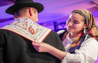 Una donna sorride mentre sistema un lecos dos namorados ad un uomo in abiti tradizionali durante un San Valentino in Portogallo. Entrambi sono vestiti con costumi folcloristici vivaci, con l'uomo che indossa un cappello nero e una giacca, evocando lo spirito di questo vivace evento culturale.