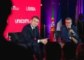 Carlos Moedas e Emmanuel Macron in giacca e cravatta sono seduti sul palco dell' unicorn factory di Lisbona durante un evento. Stanno parlando nei microfoni. Dietro di loro ci sono bandiere portoghesi e francesi, che catturano l'essenza del dibattito in corso. Le sagome del pubblico incombono in primo piano.