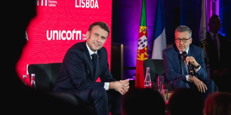 Carlos Moedas e Emmanuel Macron in giacca e cravatta sono seduti sul palco dell' unicorn factory di Lisbona durante un evento. Stanno parlando nei microfoni. Dietro di loro ci sono bandiere portoghesi e francesi, che catturano l'essenza del dibattito in corso. Le sagome del pubblico incombono in primo piano.