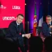 Carlos Moedas e Emmanuel Macron in giacca e cravatta sono seduti sul palco dell' unicorn factory di Lisbona durante un evento. Stanno parlando nei microfoni. Dietro di loro ci sono bandiere portoghesi e francesi, che catturano l'essenza del dibattito in corso. Le sagome del pubblico incombono in primo piano.