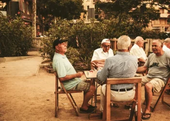 Cinque uomini anziani sono seduti all'aperto attorno a un tavolino, impegnati a conversare e a condividere consigli di longevità. Sono vestiti in modo casual, con alberi ed edifici sullo sfondo. L'ambiente sembra rilassato e cordiale.