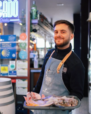 Un sorridente ristoratore che indossa un grembiule regge un vassoio di pesce fresco nrl mercato di Matosinhos. Sullo sfondo, una luminosa insegna al neon illumina, ricordando calda atmosfera.