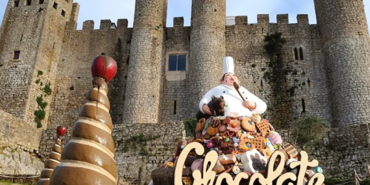 Una grande scultura a tema cioccolato raffigurante uno chef e vari dolciumi in presentazione del Festival del Cioccolato di Obidos, sullo sfondo delle imponenti mura in pietra del castello storico. In primo piano, strutture a cono di cioccolato accompagnano un cartello con la scritta Cioccolato.