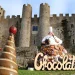 Una grande scultura a tema cioccolato raffigurante uno chef e vari dolciumi in presentazione del Festival del Cioccolato di Obidos, sullo sfondo delle imponenti mura in pietra del castello storico. In primo piano, strutture a cono di cioccolato accompagnano un cartello con la scritta Cioccolato.
