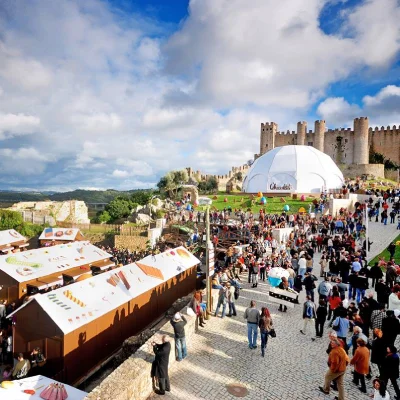 Il vivace Festival del Cioccolato di Obidos panoramica all'aperto di fronte al castello medievale, con le bancarelle marroni e la grande tenda a cupola bianca. La strada acciottolata è piena di persone che si godono l'evento sotto un cielo parzialmente nuvoloso, offrendo una deliziosa panoramica del borgo.