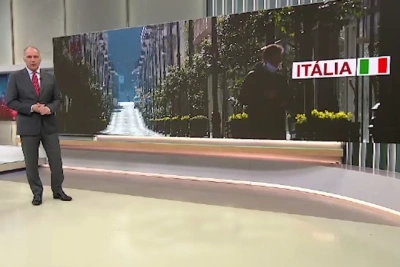 Nella Giornata in memoria delle vittime del Covid un uomo in piendi all'interno dello studio del tg in Portogallo parla con le mani giunte. Si vede un maxi schermo con l'immagine di una strada deserta in Italia con alberi, palazzi e il riflesso del sole.