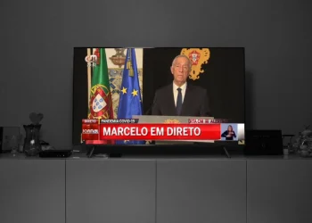 Nella Giornata in memoria delle vittime del Covid in Portogallo un televisore trasmette un servizio del Tg in cui compare il Presidente della repubblica Macelo Rebelo de Sousa, accanto alle bandiere del Portogallo e dell'UE. Sullo schermo si legge "Pandemia COVID-19" e "Marcelo em direto". È presente un interprete del linguaggio dei segni.
