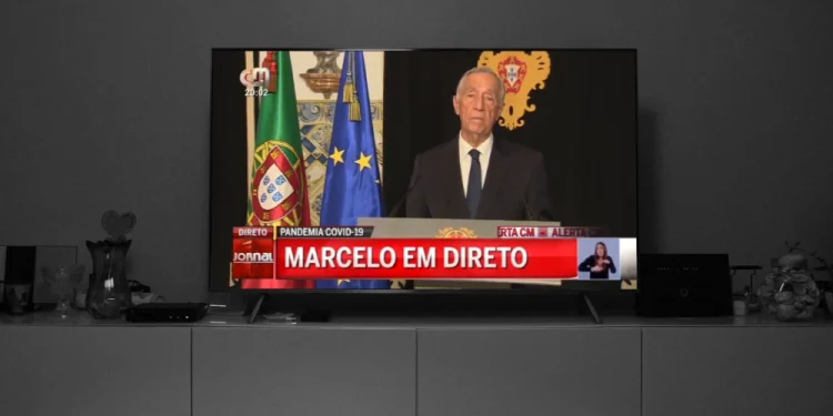 Nella Giornata in memoria delle vittime del Covid in Portogallo un televisore trasmette un servizio del Tg in cui compare il Presidente della repubblica Macelo Rebelo de Sousa, accanto alle bandiere del Portogallo e dell'UE. Sullo schermo si legge "Pandemia COVID-19" e "Marcelo em direto". È presente un interprete del linguaggio dei segni.