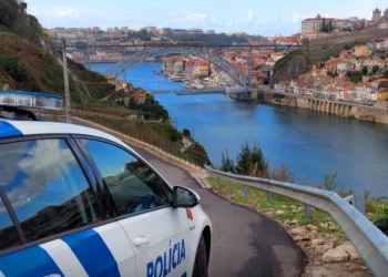 Un'auto della polizia è parcheggiata su una strada che si affaccia sulla città di Porto, come descritto nel Rapporto Sicurezza Interna 2024. Il ponte attraversa la città con edifici colorati sulla collina. Sulla fiancata dell'auto c'è scritto "Polícia" e il cielo è parzialmente nuvoloso.