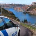 Un'auto della polizia è parcheggiata su una strada che si affaccia sulla città di Porto, come descritto nel Rapporto Sicurezza Interna 2024. Il ponte attraversa la città con edifici colorati sulla collina. Sulla fiancata dell'auto c'è scritto "Polícia" e il cielo è parzialmente nuvoloso.