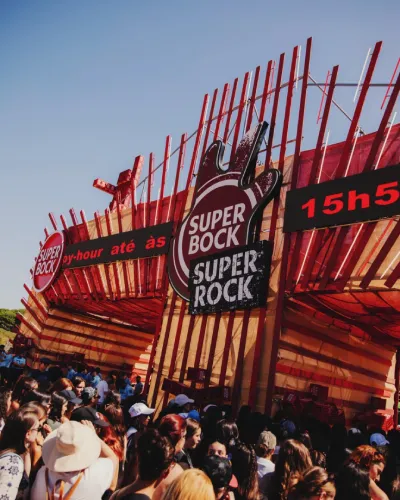 In una giornata dal cielo azzurro, una grande folla si raduna all'ingresso del Super Bock Super Rock. Una grande struttua di design di colore arancione con led colorati e la scritta  Super Bock Super Rock.
