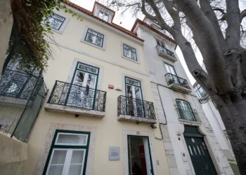Una vista esternna del nuovo centro accoglienza senzatetto di Lisbona. Edificio giallo chiaro a tre piani con finestre bordate di verde e balconi in ferro battuto. Un grande albero si erge lì vicino, ombreggiando l'ingresso del portone del piano terra.