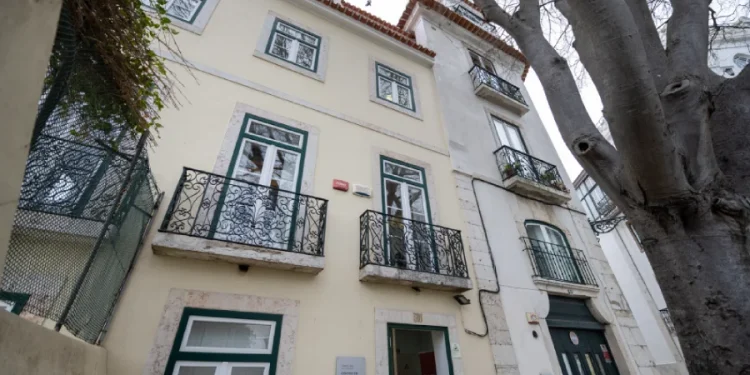 Una vista esternna del nuovo centro accoglienza senzatetto di Lisbona. Edificio giallo chiaro a tre piani con finestre bordate di verde e balconi in ferro battuto. Un grande albero si erge lì vicino, ombreggiando l'ingresso del portone del piano terra.