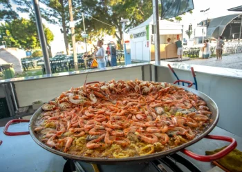 Tra gli eventi c'è una grande padella di paella di pesce che mostra gamberi e cozze in esposizioni in un festival all'aperto in Algarve. Sullo sfondo, ci sono tavoli, alberi e tende, che suggeriscono un'atmosfera festosa piena dei sapori vibranti.