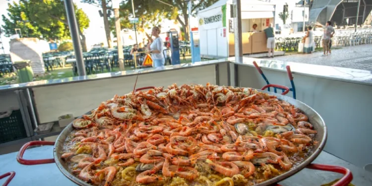 Tra gli eventi c'è una grande padella di paella di pesce che mostra gamberi e cozze in esposizioni in un festival all'aperto in Algarve. Sullo sfondo, ci sono tavoli, alberi e tende, che suggeriscono un'atmosfera festosa piena dei sapori vibranti.