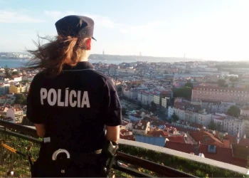 Un agente di polizia donna con i capelli lunghi è in piedi su un punto panoramico, vigile per i segnali di furti appartamento a Lisbona in Portogallo. La parola "Policia" è visibile sul retro dell'uniforme in mezzo alla vasta distesa di edifici, il fiume Tejo sotto il cielo limpido.