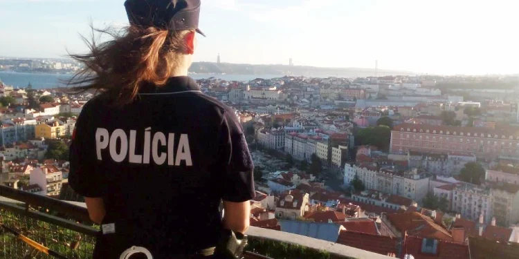 Un agente di polizia donna con i capelli lunghi è in piedi su un punto panoramico, vigile per i segnali di furti appartamento a Lisbona in Portogallo. La parola "Policia" è visibile sul retro dell'uniforme in mezzo alla vasta distesa di edifici, il fiume Tejo sotto il cielo limpido.