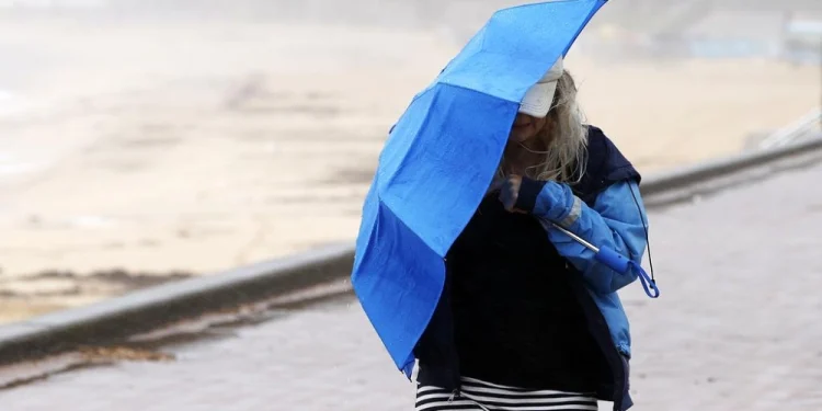Una persona con una giacca scura e pantaloni a righe lotta per tenere un ombrello blu contro il vento forte su un sentiero lastricato vicino alla spiaggia sabbiosa, tipico del meteo Portogallo. Il tempo è nuvoloso e nebbioso.