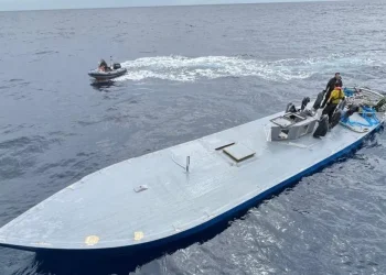 Nell'oceano: un piccolo motoscafo con due persone sullo sfondo e un sottomarino più grande, a ponte piatto, sequestrato per trafficare di droga con tre persone in primo piano. L'imbarcazione più grande sembra avere delle attrezzature sul ponte. Il mare è calmo e il cielo è coperto.