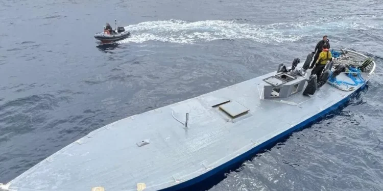 Nell'oceano: un piccolo motoscafo con due persone sullo sfondo e un sottomarino più grande, a ponte piatto, sequestrato per trafficare di droga con tre persone in primo piano. L'imbarcazione più grande sembra avere delle attrezzature sul ponte. Il mare è calmo e il cielo è coperto.