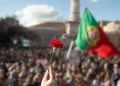 In primo piano una mano tiene un garofano rosso, sullo sfondo una grande folla per la Sfilata per il 25 aprile a Lisbona, con la bandiera portoghese che sventola.