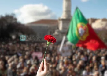In primo piano una mano tiene un garofano rosso, sullo sfondo una grande folla per la Sfilata per il 25 aprile a Lisbona, con la bandiera portoghese che sventola.