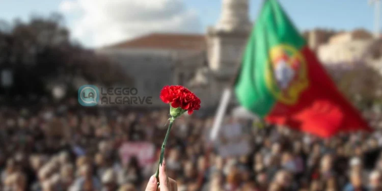 In primo piano una mano tiene un garofano rosso, sullo sfondo una grande folla per la Sfilata per il 25 aprile a Lisbona, con la bandiera portoghese che sventola.