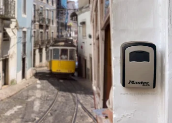 Un tram giallo percorre una ripida e stretta strada acciottolata di Lisbona. In primo piano, un lucchetto "Master Lock" – di norme utilizzato per Airbnb e Affitti Brevi – è appeso a una parete bianca.