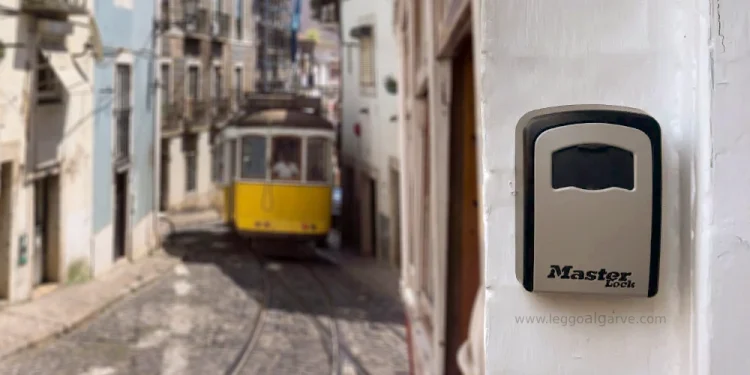 Un tram giallo percorre una ripida e stretta strada acciottolata di Lisbona. In primo piano, un lucchetto "Master Lock" – di norme utilizzato per Airbnb e Affitti Brevi – è appeso a una parete bianca.